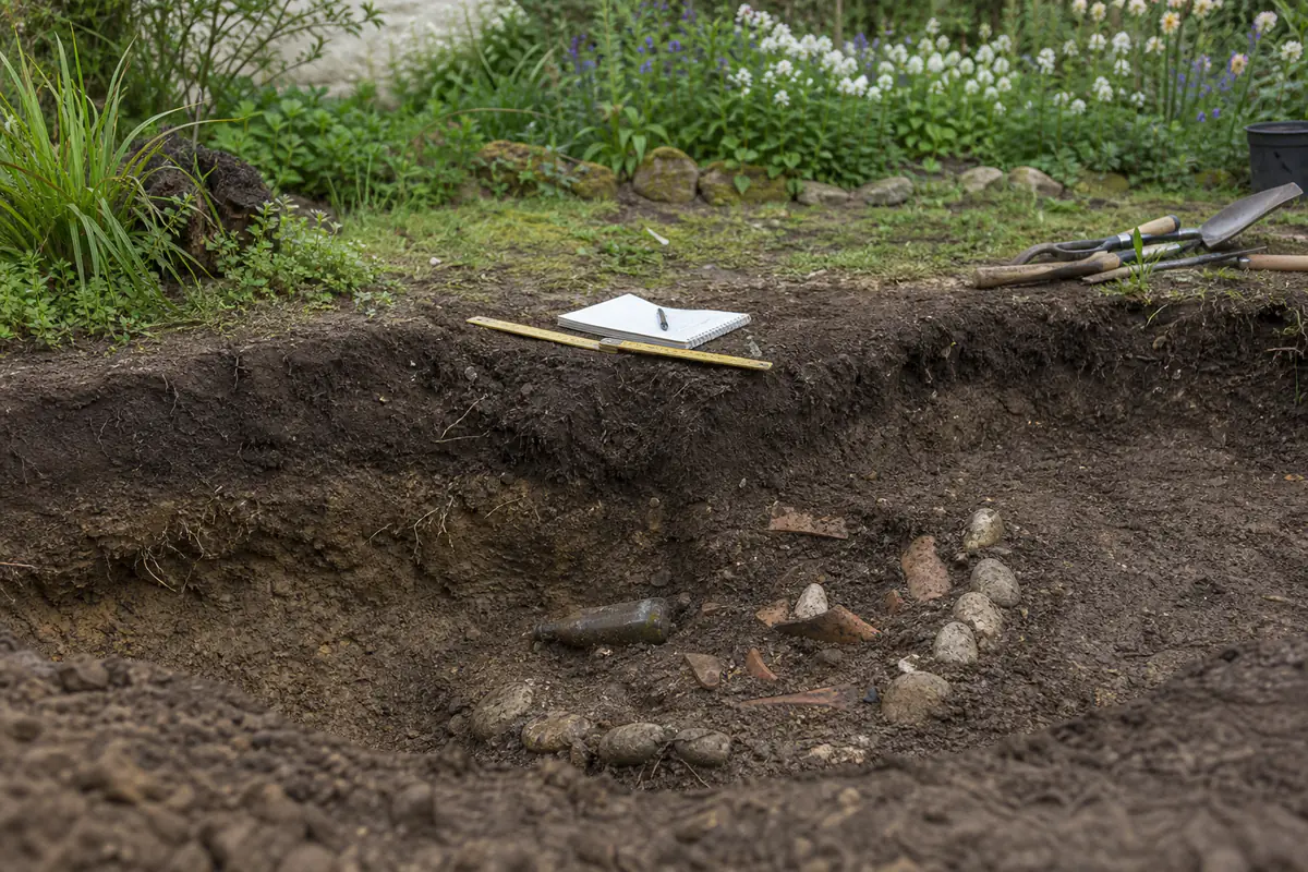 Tessons de poterie, bouteille ancienne et pierres visibles dans une tranchée de jardin avant leur relevé